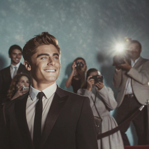 Zac Efron, in black suit and tie, smiling at glamorous awards night. Crowds of celebrities and flashing cameras.