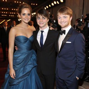 Harry Potter cast reunion. Emma Watson in a flowing blue gown, Daniel Radcliffe in a classic black tuxedo, and Rupert Grint in a navy suit. Glittering indoor event, surrounded by photographers and flashing lights.