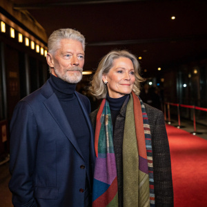 Stephen Lang in a navy suit and Kristina Watson in a multicolored scarf and turtleneck at a film festival premiere. They stand beside a dimly lit red carpet inside a theater.