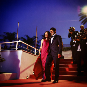 Lara Pulver and Raza Jaffrey at a film festival. Lara in a flowing red dress. Raza in a black tuxedo. They are posing for photographers on the red carpet.