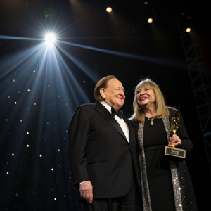 Sheldon Adelson and Miriam Ochsorn at an awards ceremony. Dark stage, spotlight on them, awards ceremony backdrop.
