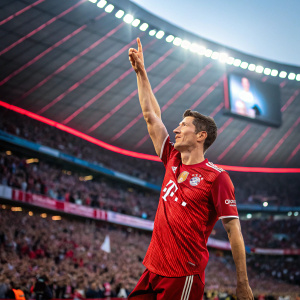 Robert Lewandowski, raising his arm in celebration, in the middle of a packed stadium, filled with cheering fans. The backdrop showcases the vibrant Allianz Arena.
