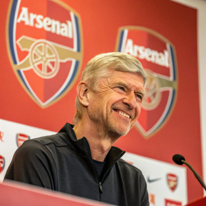 Arsène Wenger, with a kind smile and slight beard, stands at a press conference. Arsenal FC logo backdrop