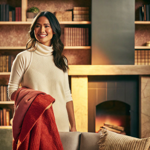 Olivia Munn, Asian actress with a friendly smile, standing in a cozy living room with bookshelves and a fireplace in the background. She wears a white turtleneck and a cozy sweater. Warm lighting.