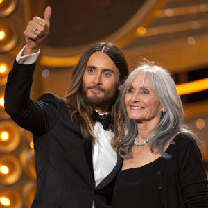 Jared Leto and Constance Leto at an awards show. Jared giving a thumbs-up, Constance with a gentle smile. Warm, golden-hued background.