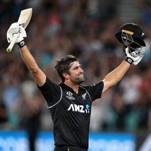 Colin de Grandhomme, cricket player, celebrates victory. Dynamic pose with arms raised. Black jersey with sponsor logos. Blurred stadium crowd in background.