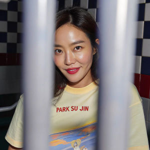 Close-up of Park Su-jin, Korean actress. Wearing yellow t-shirt with graphic and text. Warm smile. Checkerboard red and white background