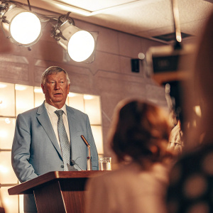 Sir Alex Ferguson, wearing a blue suit and tie, at a press conference. Bright studio lights, modern backdrop with company logos.