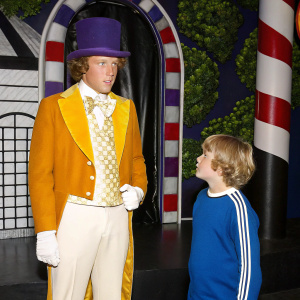 Gene Wilder as Willy Wonka standing in the chocolate factory. Peter Ostrum, young protagonist, looks on. Lush greenery and whimsical decor surround them. Photo from the movie