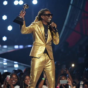Lil Wayne, wearing a shiny gold suit, is at the BET Awards show, holding a trophy and addressing the audience.