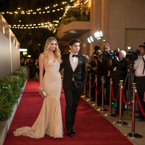 Veronica Dunne and Max Ehrich at a gala event. Veronica in a stunning beige gown, Max in a black tuxedo. Red carpet ambiance with flashing cameras.