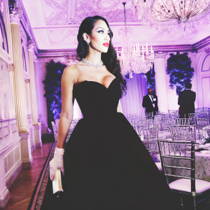 Erica Mena at a gala event. She's in a black dress with red lipstick, standing in a room with gold and blue decor.