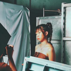 Young Asian girl, with her hair in pigtails, wearing a brown strapless top. She is focused intently on a task in a neutral gray studio setting.