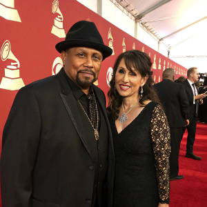 Aaron Neville and Sarah A. Friedman attending the Grammy Awards. Black fedora, dark hair, red carpet, formal attire, red backdrop with Grammy logos, other celebrities present.