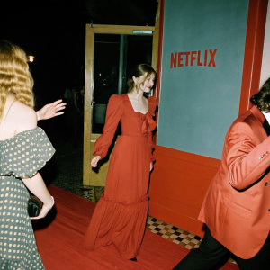 Celebrities and guests at the Netflix Enola Holmes 2 premiere. They are dressed in elegant evening wear. A bright, lively atmosphere with a Netflix-branded backdrop behind them.