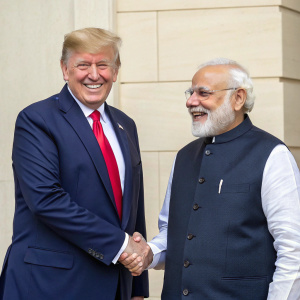 President Donald Trump and Prime Minister Narendra Modi sharing a handshake. The backdrop is a simple, neutral-colored wall. Trump, in a navy suit and red tie, stands next to Modi, dressed in a traditional Indian sherwani.
