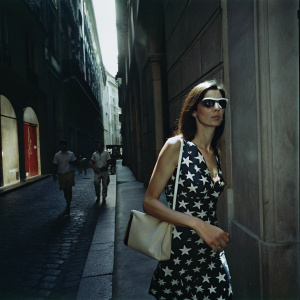 Famke Janssen strolls down a bustling Parisian street, framed against the backdrop of a quaint, cobblestone alleyway. Dressed in a sleeveless, star-patterned dress and carrying a structured handbag, she moves with a relaxed elegance, sunglasses shielding her eyes from the sun.