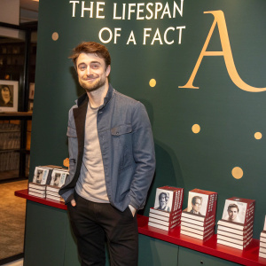 Daniel Radcliffe in a casual outfit, standing at the book launch of 'The Lifespan of a Fact'. The backdrop features the title and publisher logos in a bold, modern design.