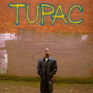 Photograph of Tupac Shakur. Wearing a classic black leather jacket, standing in front of graffiti-covered brick wall. Name 'Tupac' in bright, vibrant colors