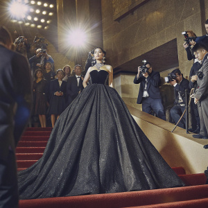 Danna Paola at a glitzy awards ceremony. She is in a floor-length, black sequined gown and bold jewelry. Crowded backdrop of flashing cameras and celebrities.