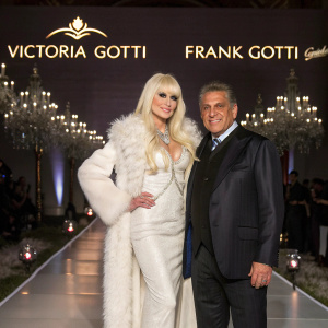 Victoria Gotti and Frank Gotti Agnello posing at a glamorous fashion show. The backdrop features a chic designer logo. The setting is an upscale event with elegant decor.
