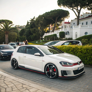White Volkswagen Golf GTI W12 650 Concept car parked outdoors, among other vehicles. Sleek design with white body and black and red accents. Lush greenery and white buildings in background. Daytime natural light