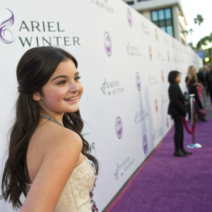 Ariel Winter, young actress, smiling in a red carpet event. Purple carpet, with sponsors logos and text on white wall backdrop.
