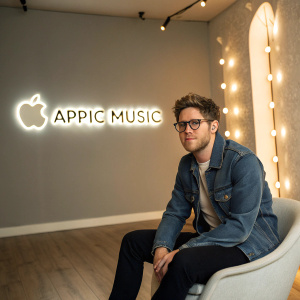 Niall Horan at the Apple Music studio, sitting in a modern, minimalist room. He's wearing a stylish denim jacket and glasses. Surrounded by glowing lights and a large streaming service logo