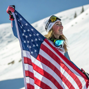 Mikaela Shiffrin, with an ecstatic smile, stands proudly in her ski gear beside the American flag. The backdrop is a snowy mountain landscape. The flag's red and white stripes contrast vividly against the snow.