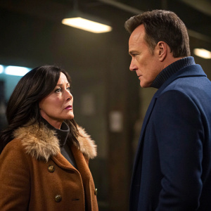 Shannen Doherty and Julian McMahon in a tense scene from a drama series. They both have serious expressions. Shannen is in a brown coat with fur trim, Julian in a navy blazer. Shot in a dimly lit room with a blurred background