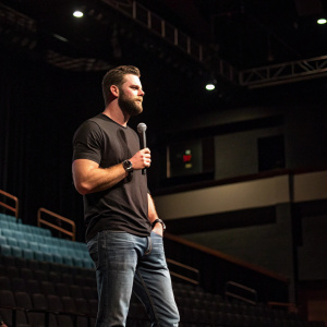 Brandon Belt at a press conference for his new book, dressed in a casual black t-shirt and jeans. Empty stage with dim lighting.