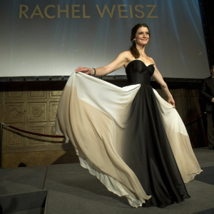 Rachel Weisz at an awards ceremony. Elegant black and white dress with strapless neckline. White fabric flowing in the breeze. White backdrop with repeating gold logos.