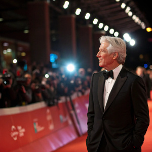 Richard Gere, elegantly dressed in a black tuxedo, at the premiere of a new film. Flashing lights and a bustling crowd in the background.