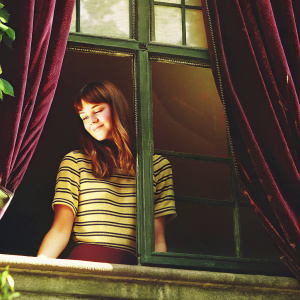 Hailee Steinfeld, fair-skinned and smiling gently. Wearing a casual striped top. Rich blue backdrop.