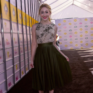 Chloe Lukasiak, radiant at the Teen Choice Awards, wearing a floral top and green skirt. She beams against a vibrant yellow backdrop adorned with event logos and banners.