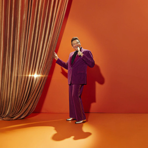 Cristiano Malgioglio, in a purple suit, on stage at a gala event. Shimmering backdrop and dramatic lighting.