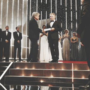Roman Polanski receiving an award from Harrison Ford at a glitzy gala. Ford in a black suit, Polanski in a tuxedo. Bright stage lights, red carpet ambiance.
