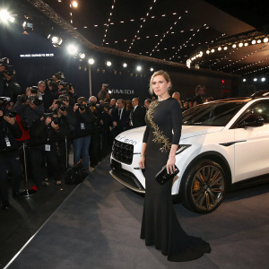 Kate Winslet attends the launch event of a luxury car brand. Wearing a sleek, long black dress with subtle gold embroidery. Crowded venue with many photographers and onlookers.