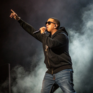 Daddy Yankee on the stage, wearing a black hoodie and jeans. Poses energetically with a microphone. Dark, smoky background