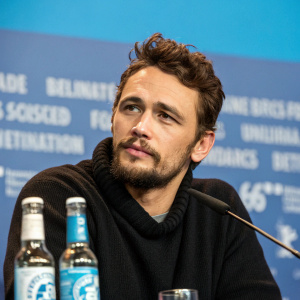 James Franco, caucasian, bearded, focused expression. Wearing black sweater. Solid blue background with text elements. Press conference at Berlinale International Film Festival. Microphone in front of him. Beverage bottles on table.