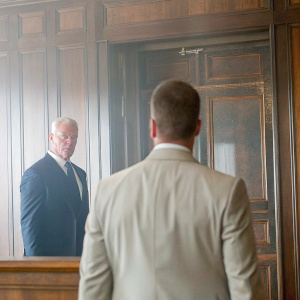 Neal McDonough as Sean Cahill in a tense office scene from 'Suits'. He wears a dark suit and stands in a room with dark wooden panels, the other actor facing away from the camera.
