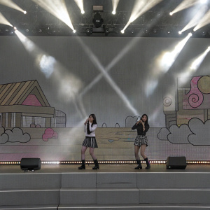 Yujue Sinb and Sowon from GFRIEND performing on a brightly lit stage. Colorful, animated backdrop