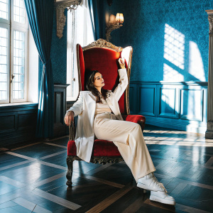 Mallory Jansen, as Madalena, sits elegantly on a red velvet throne. Dark wooden floor, illuminated by flickering torches on the walls.
