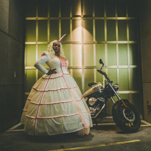Sheila Bomb, character with a unicorn-like horn and a bald head, wears a voluminous white dress with a crinoline skirt and pink accents. Standing confidently next to a futuristic motorcycle on a metallic grey grid surface.