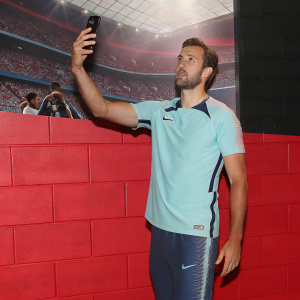 Harry Kane, in a Nike jersey, taking a selfie at the stadium. Fans and photographers in background.