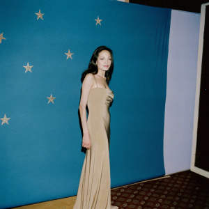 Angelina Jolie at BAFTA Awards, dressed in an elegant olive-green gown. She poses confidently in front of a dimly lit, star-studded backdrop.