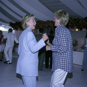 Hillary Clinton and Ellen DeGeneres laughing and chatting at a lively fundraising gala. Hillary is in a blue suit while Ellen is in a plaid shirt. Festive backdrop