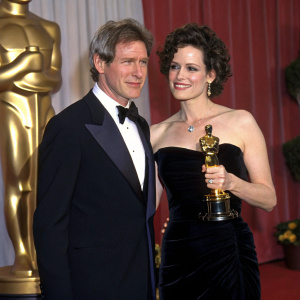 Harrison Ford and Sigourney Weaver at the Academy Awards. Dark suit and black velvet gown, holding golden trophy.
