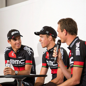 Richie Porte, Rohan Dennis, and Tejay van Garderen from BMC Racing Team at a press conference. They are wearing black and white cycling gear with BMC branding. The background is a plain white wall.
