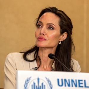 Angelina Jolie, at a UNHCR event, with serious expression, natural makeup. Neutral beige backdrop. Dark hair styled loosely.